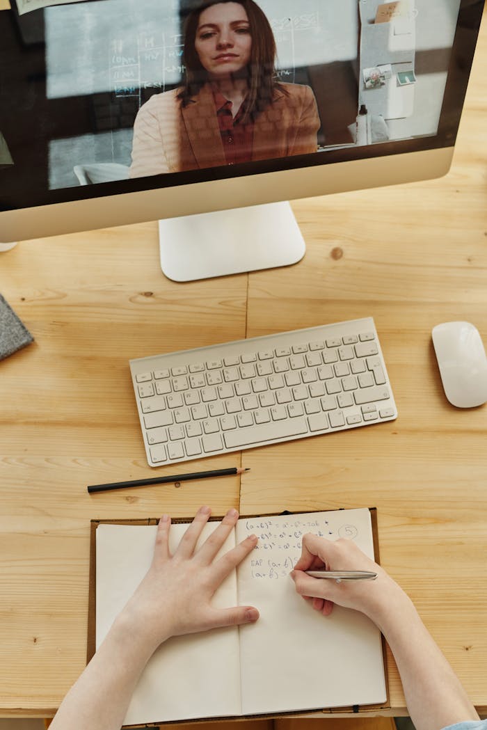 A student taking notes during an online learning session with a teacher on a monitor.